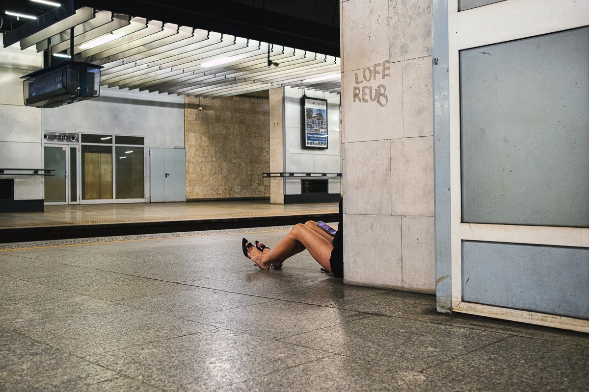 A person sitting on the floor of a train or subway station, with their legs extended out from behind a wall, holding a phone, and wearing black shoes with pointed toes.