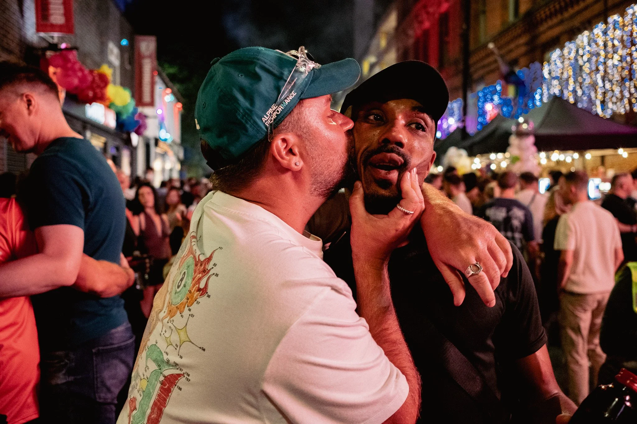 Two men sharing an affectionate moment at a lively outdoor night event. One man is kissing the other on the cheek, who appears surprised or emotional. The background features colorful lights, decorations, and a crowd of people.