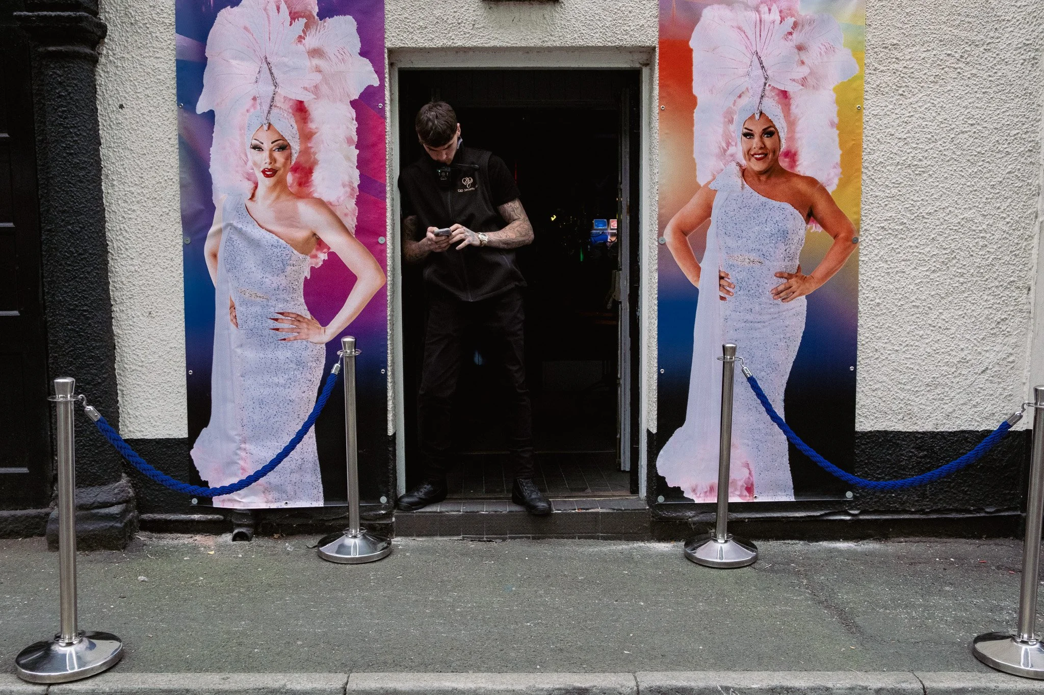 Entrance to a venue with two large posters of a woman in an extravagant costume with a feathered headdress and a sparkly dress, and a man standing in the doorway looking at his phone, surrounded by stanchions with blue ropes.