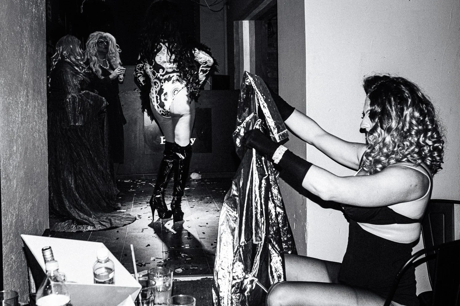 A woman with curly hair holding a shiny fabric while another woman is on a stage with performers in elaborate costumes and high heels.