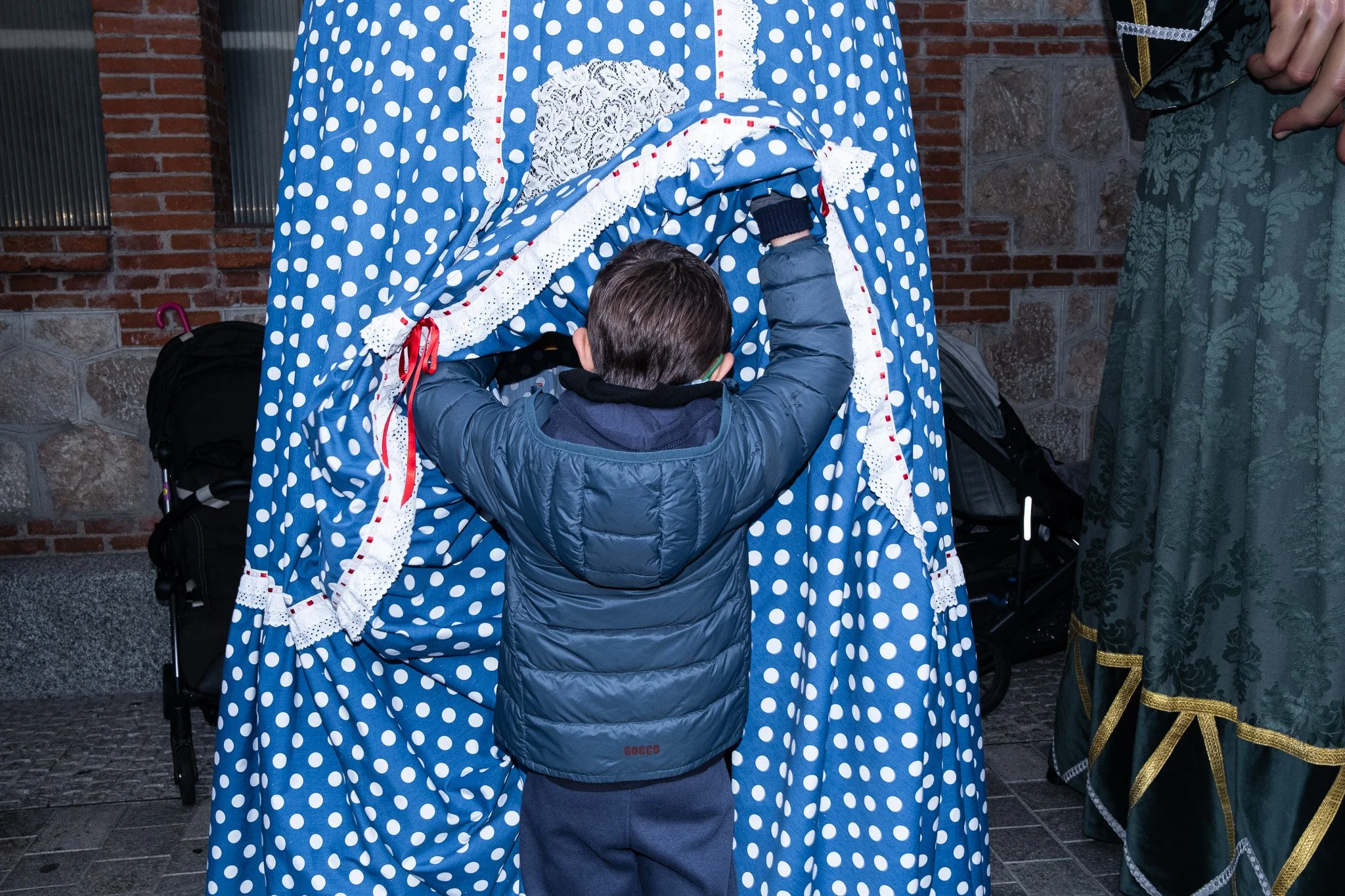 A young boy with short dark hair, wearing a blue puffer jacket, is seen from behind, venturing into a blue polka dot patterned cloth or curtain held by an adult, whose arm and part of a green embroidered garment are visible.