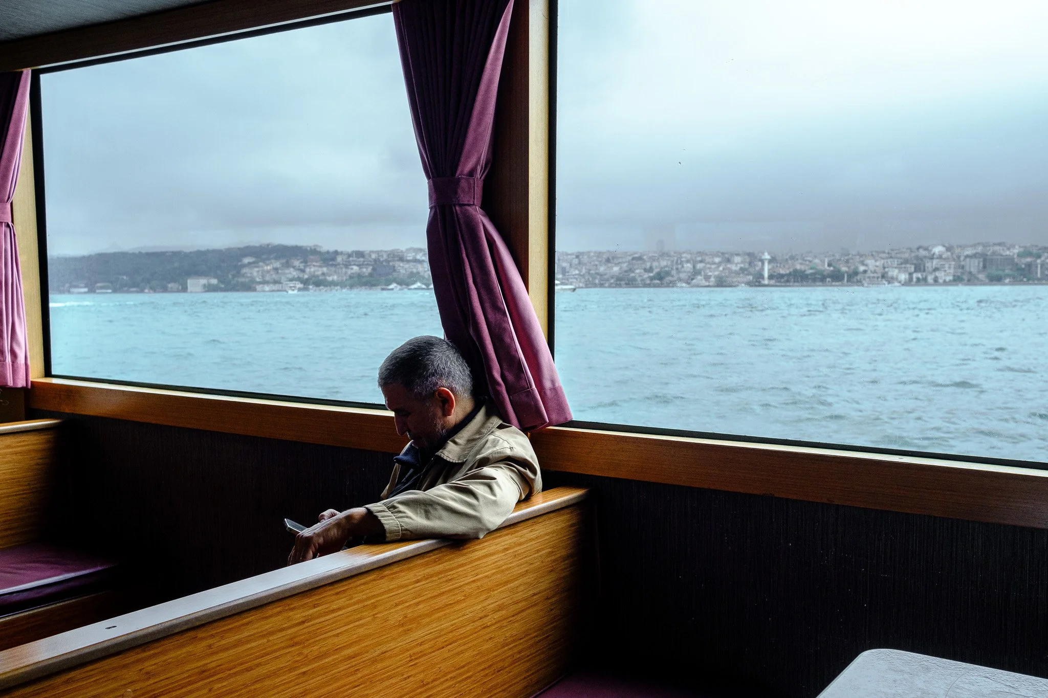 Man sitting alone on a boat, looking at his phone, with water and cityscape visible through the window behind him.