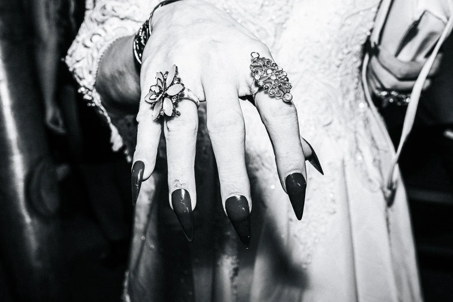 Close-up of a hand adorned with multiple rings, including a butterfly-shaped ring on the index finger and a cluster of circular rings on the middle finger, with long, pointed black nails, against a blurred background.
