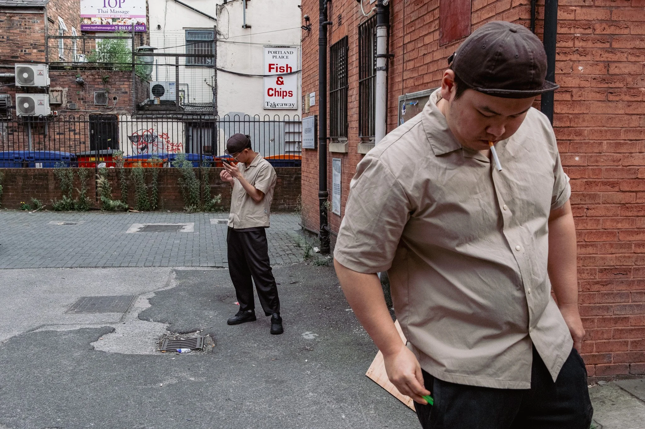 Two young men, dressed in beige shirts and black pants, stand outside near a brick wall. One is looking at his phone with a cigarette in his mouth, and the other is also looking at his phone, standing farther back.