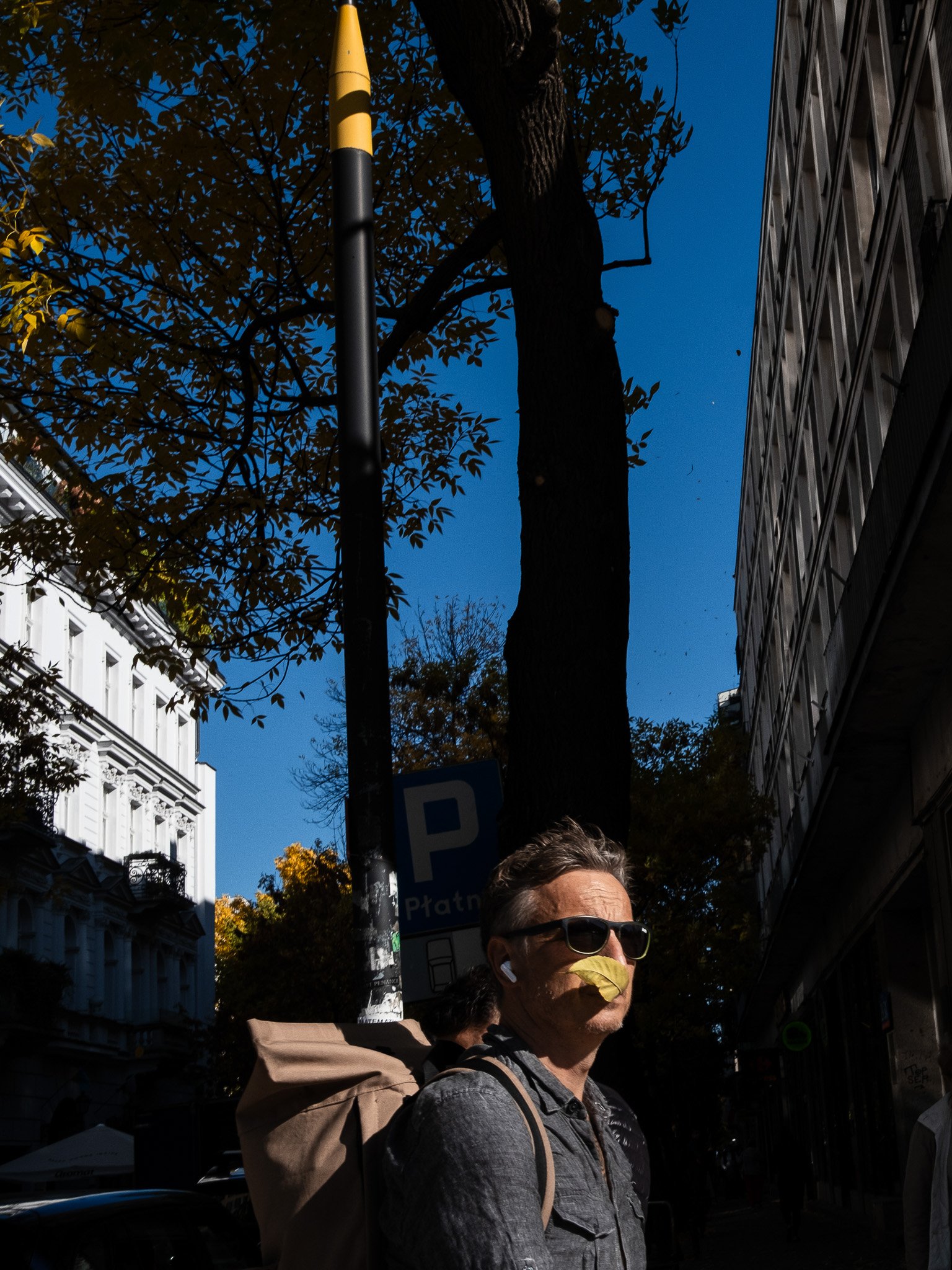 A man wearing sunglasses, a face mask, and wireless earbuds is standing outdoors on a city street with tall buildings, trees, and a parking sign visible in the background.