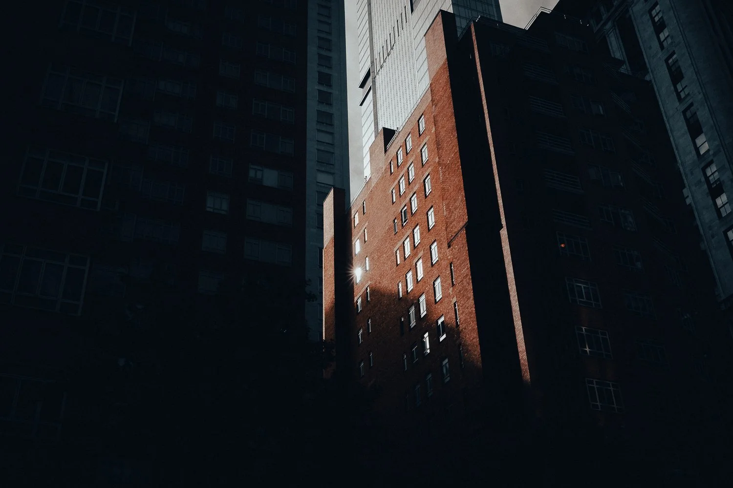 Sunlight reflects off windows of a red brick building in a city, with taller modern buildings surrounding it.