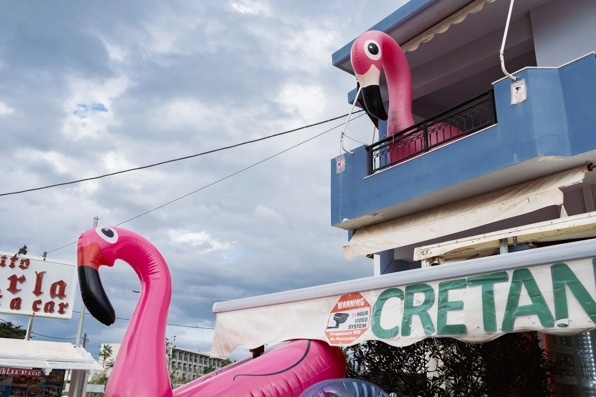 Two inflatable pink flamingo pool floats inserted into a building's balcony and a cart with a sign reading 'Cretan'.