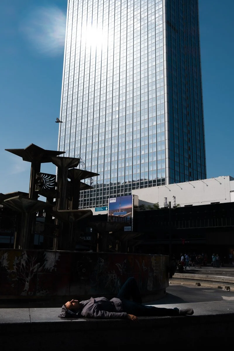 A man lying on the sidewalk in front of a tall glass skyscraper on a sunny day.