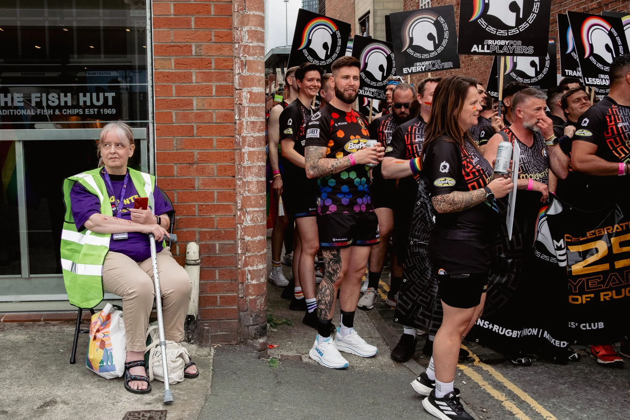 A group of people participating in a rugby event, wearing black jerseys with rainbow-colored accents, standing behind a barrier with rainbow-themed flags and banners. An older woman in a purple shirt and beige pants, sitting on a chair with a cane an