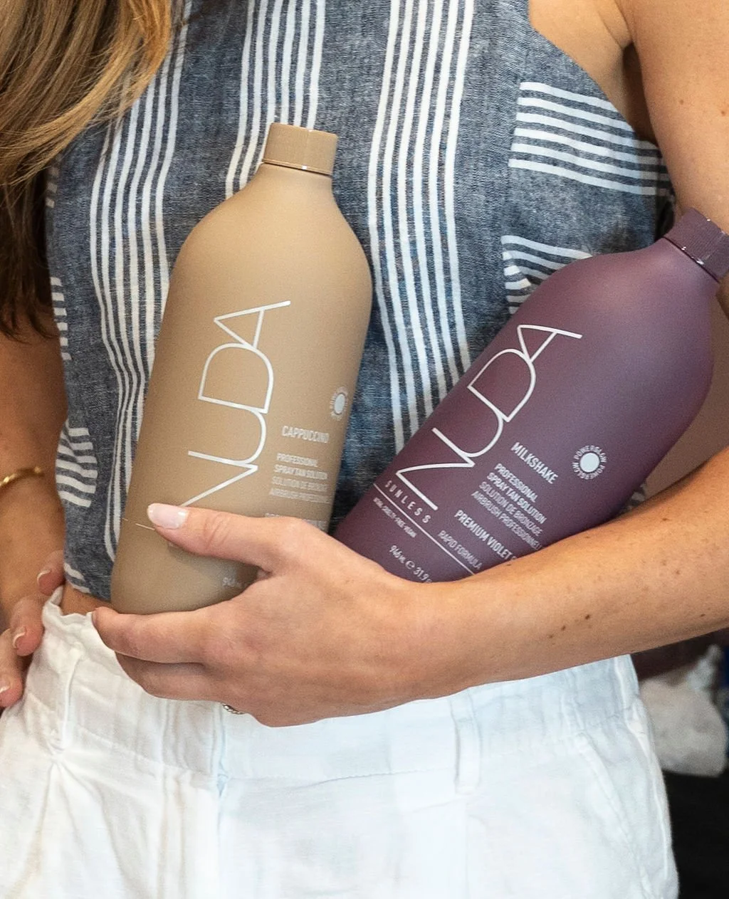 A person wearing a striped tank top and white pants is holding two large bottles of hair care products. The beige bottle is labeled 'Veda Cappuccino' and the purple bottle is labeled 'Veda Milkshake.'