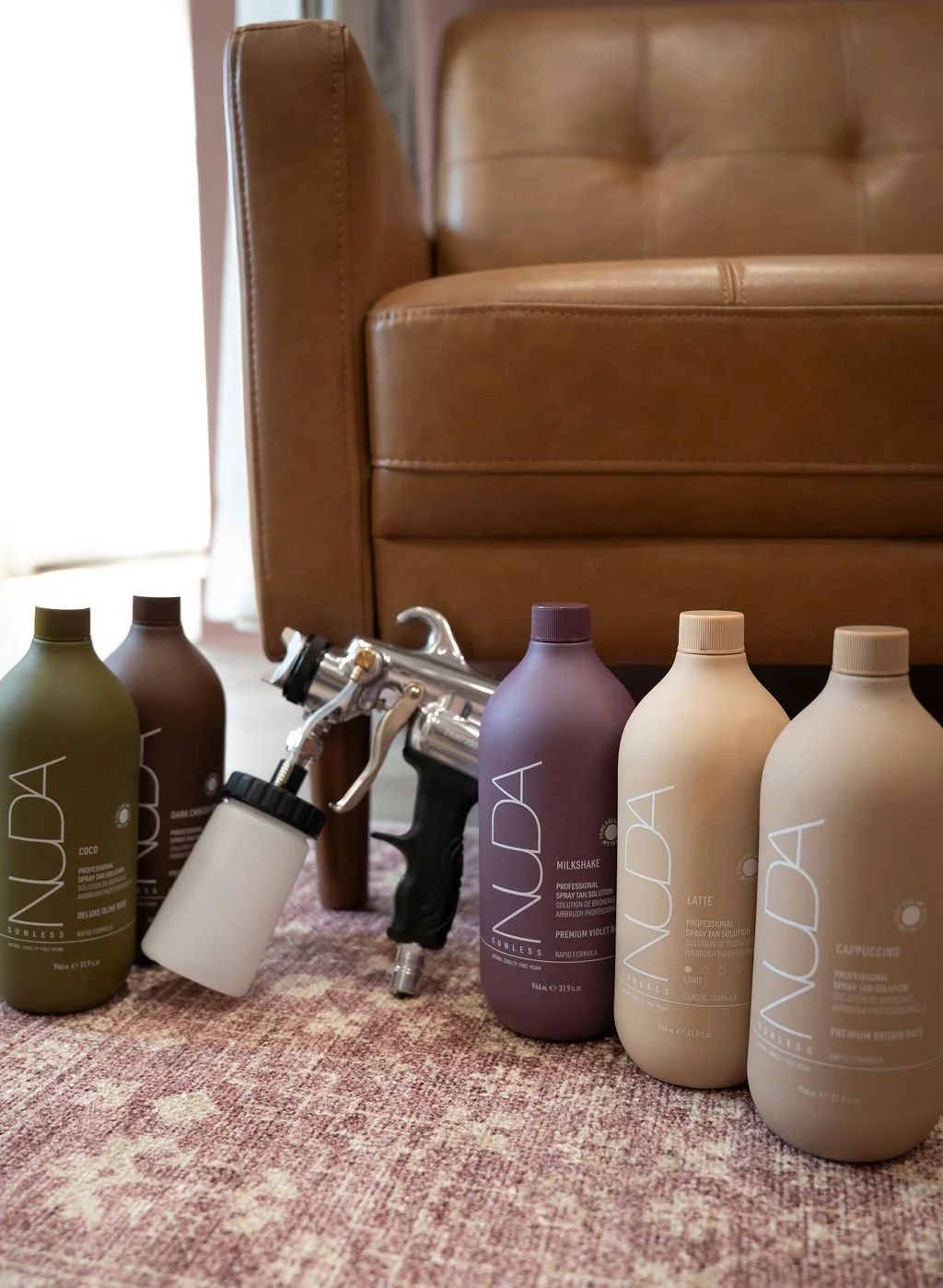 Six bottles of NUDA spray tan solution in various colors and a spray tan applicator on a pinkish carpet in front of a tan leather sofa at Tone Tans in Phoenixville.
