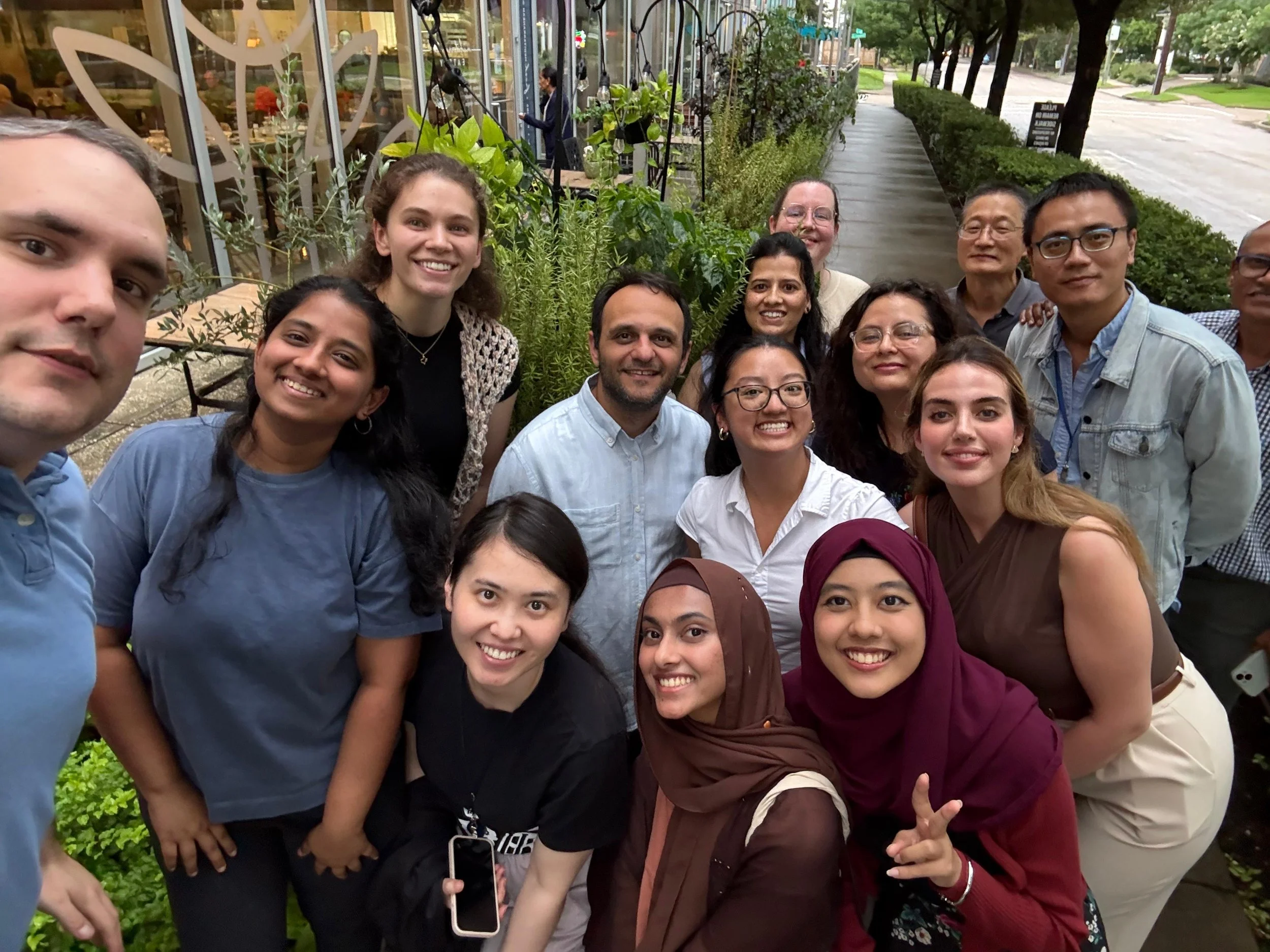Group photograph of diverse people smiling outside a restaurant, with greenery and city street in the background.
