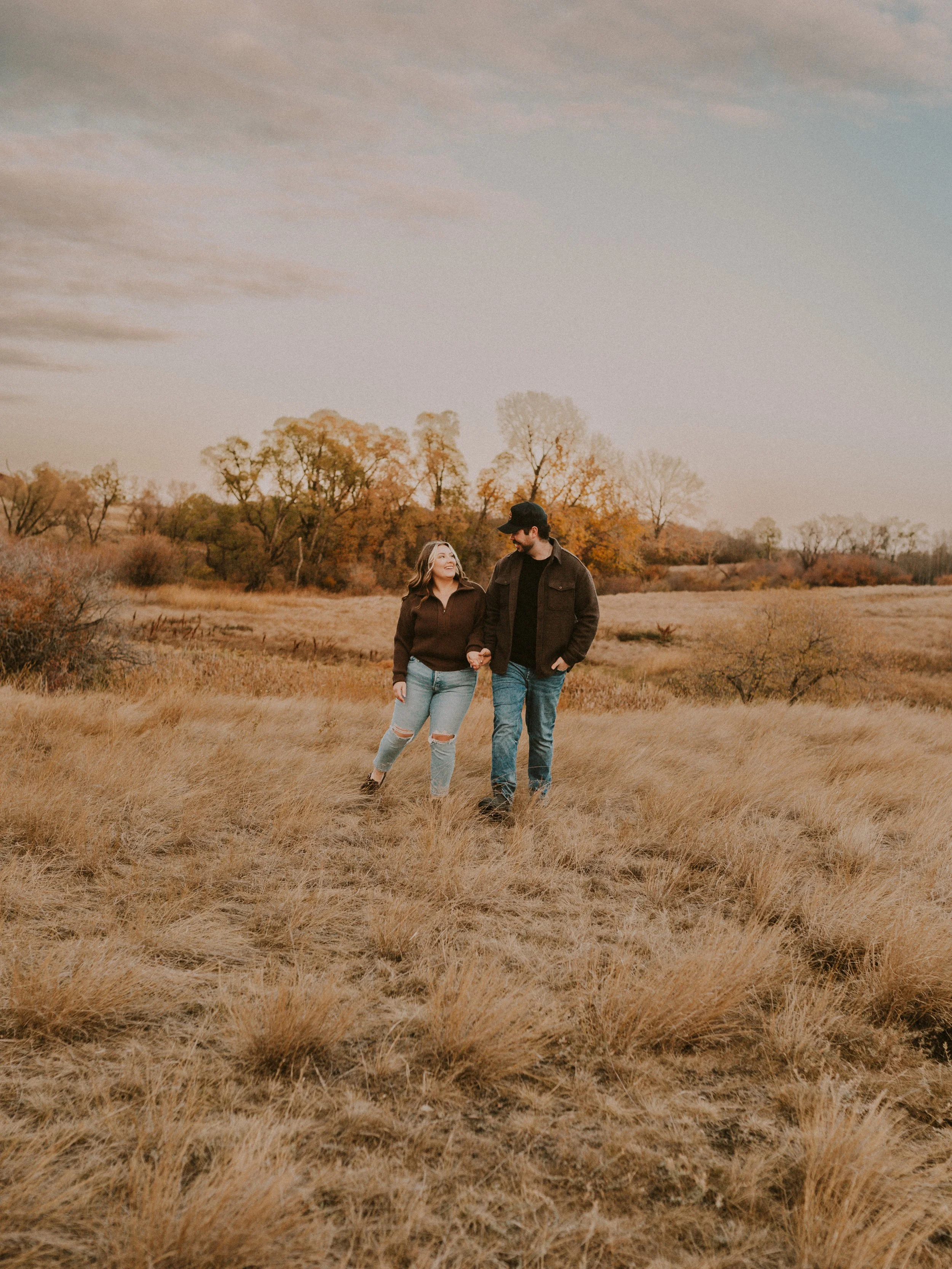 A young couple walking hand in hand through a field of tall yellow grass during autumn, with trees showing fall foliage in the background under a partly cloudy sky.