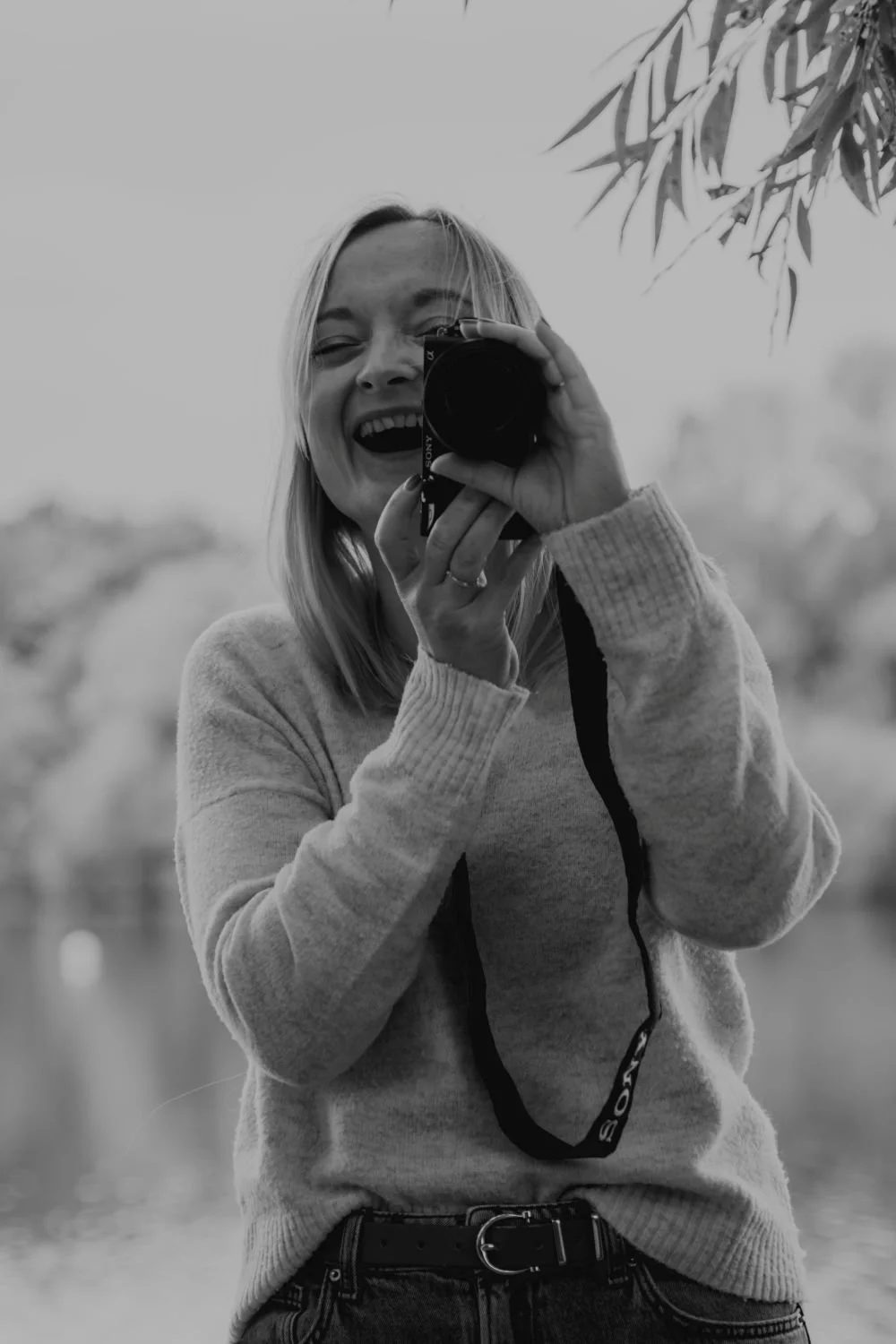 Schwarz-Weiß-Foto einer lachenden Frau, die in eine Kamera schaut, während sie ein Selbstporträt macht. Im Hintergrund sind Bäume und Wasser sichtbar.