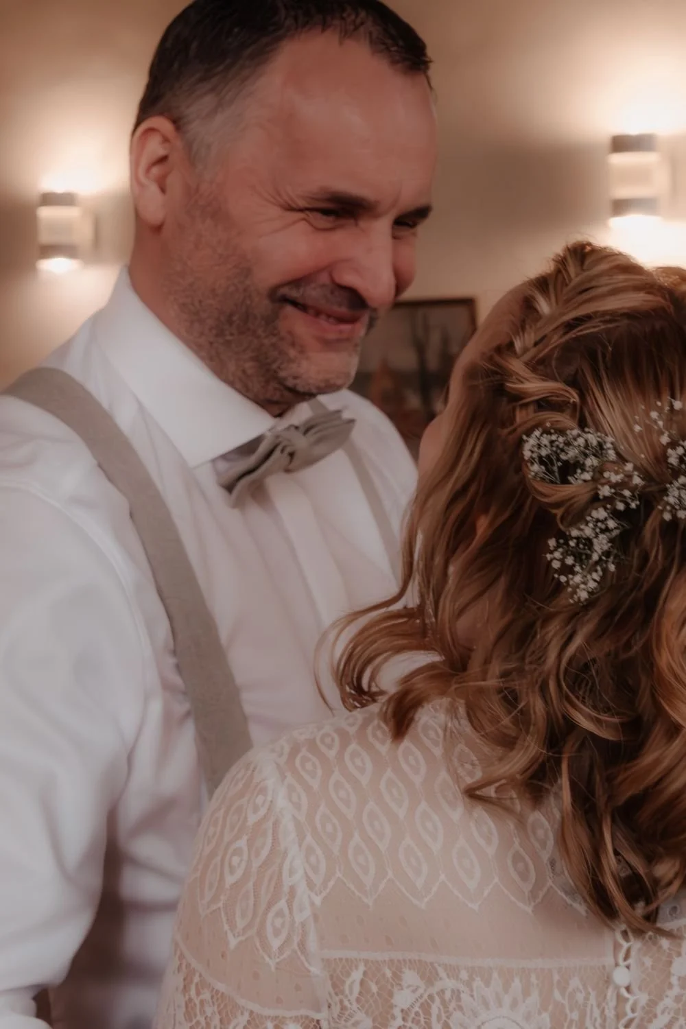 Ein Mann in einem weißen Hemd mit grauem Bolero und Schleife, lächelnd, tanzt mit einer Frau bei einer Hochzeit.