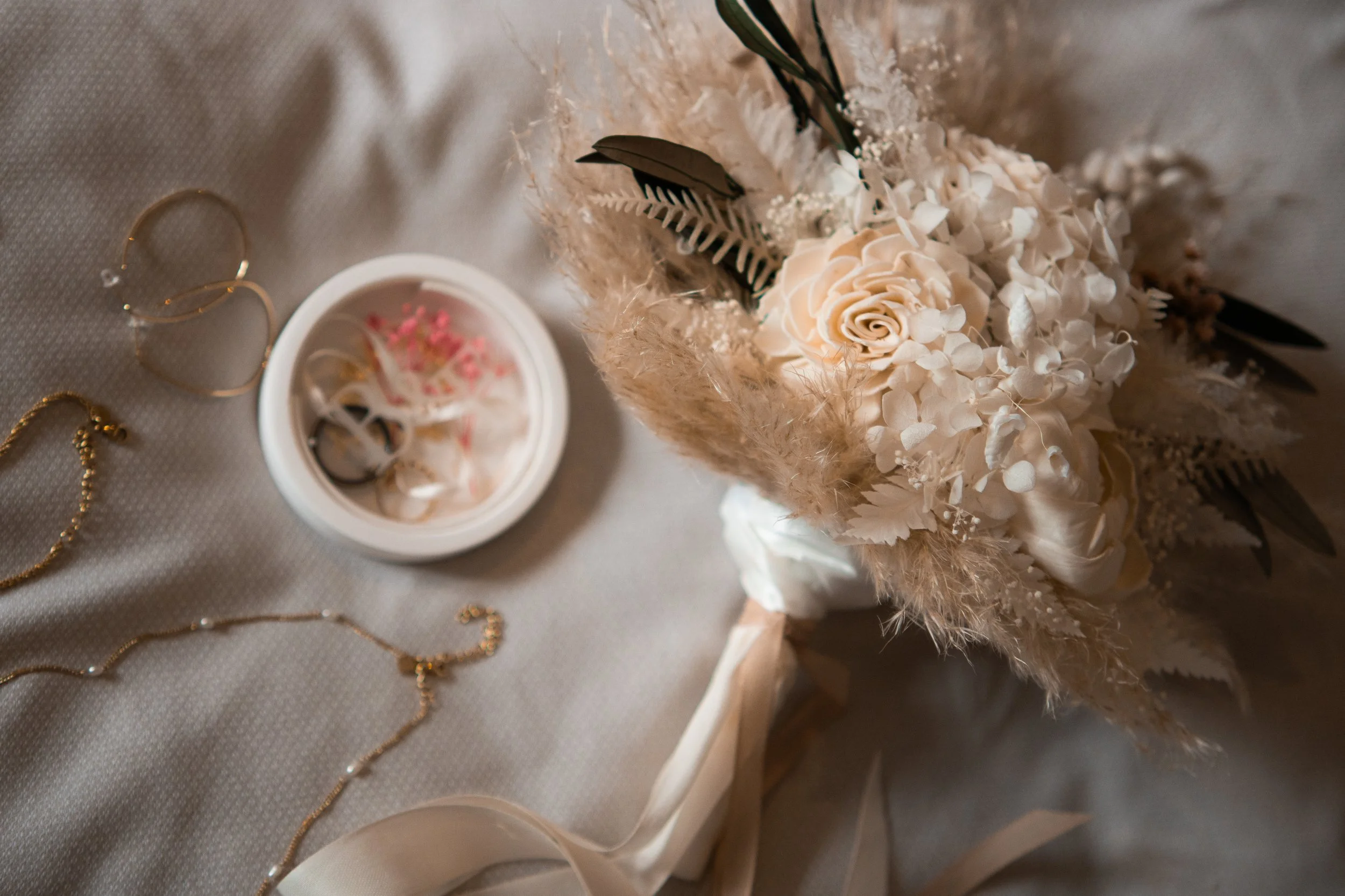 Eine Brautstrauß mit weißen Blumen, umgeben von goldenen Schmuckstücken und einem kleinen weißen Behälter auf einem hellen Stoff.