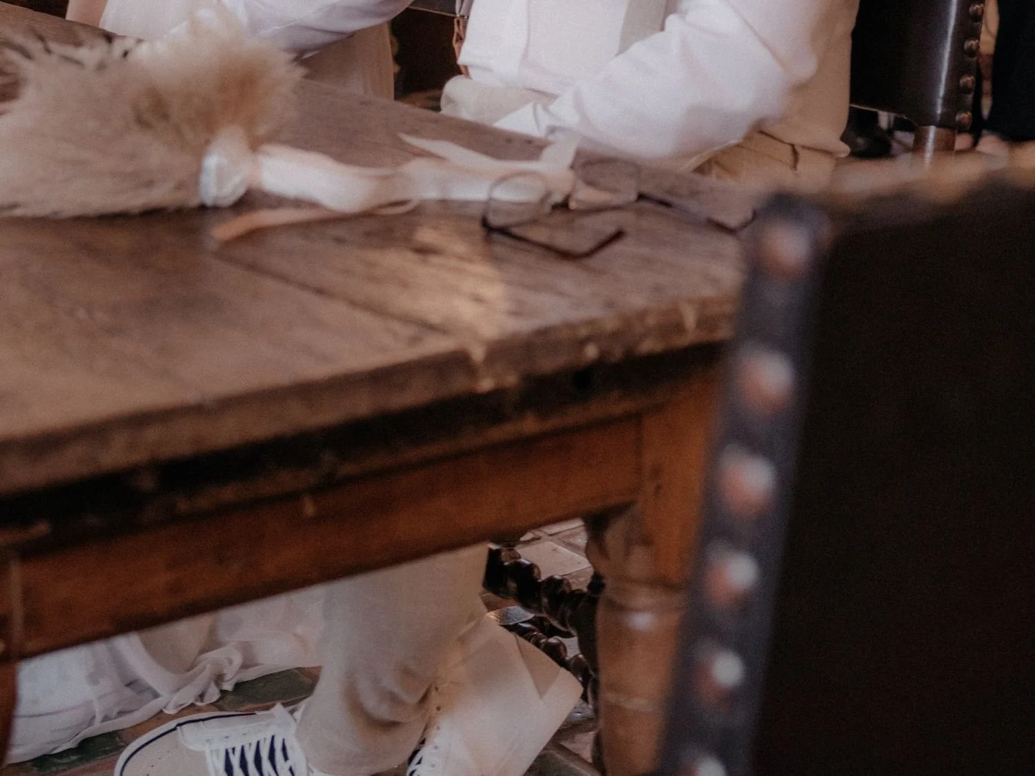 Ein Holztisch mit einem Buch, einer Brille und einem weißen Tuch. Im Hintergrund sind eine Person in weißer Kleidung und ein Teppich. Auf dem Tisch liegt eine Katze.