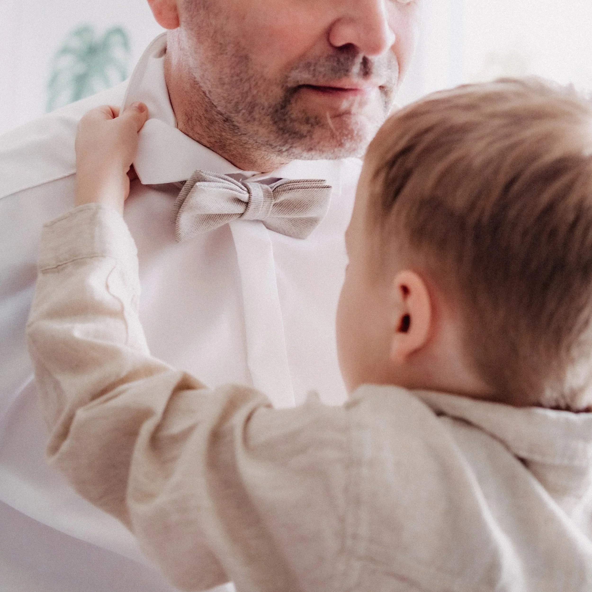 Vater und Sohn, nah beieinander, lachen und schauen sich liebevoll an, Vater trägt eine weiße Hemd mit grauem Fliege, Sohn trägt ein beige Hemd.