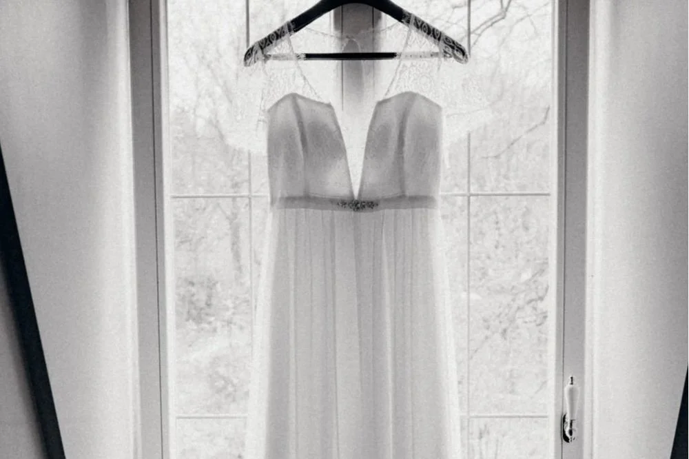Weiße Brautkleid hängt an einem Kleiderhaken vor einer Fensterscheibe.