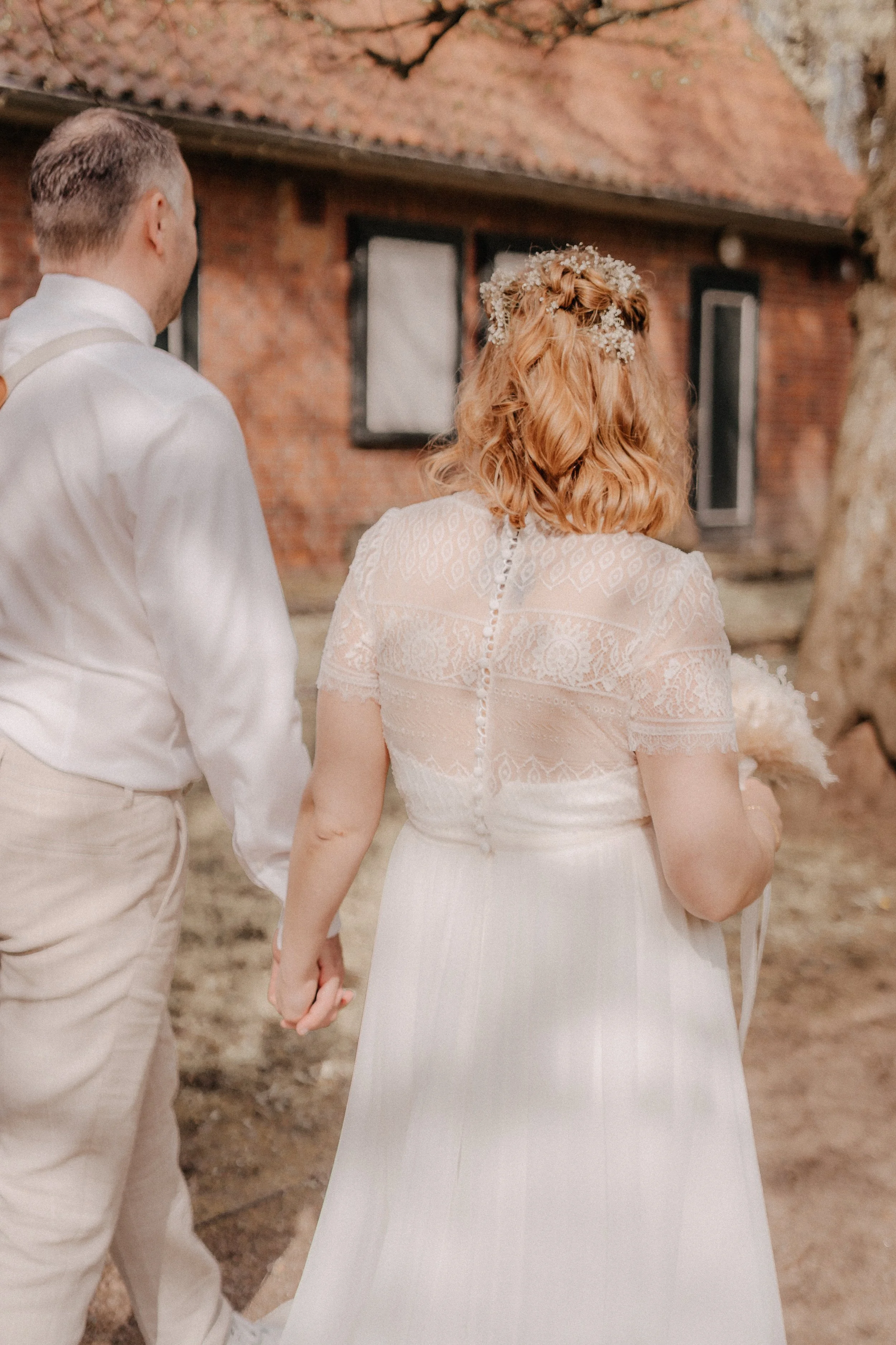 Ein Brautpaar bei einer Hochzeit, das Händchen haltend in einem Außenbereich mit Baum und einer roten Backsteinmauer im Hintergrund.