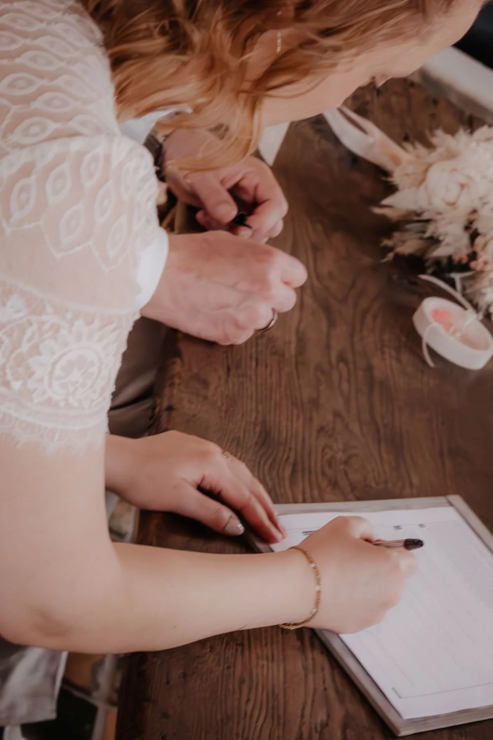 Eine ältere Frau mit blonden Haaren, die ein Dokument unterschreibt, und eine jüngere Person, die neben ihr sitzt. Es liegen Blumen und ein kleiner Korb auf dem Tisch.