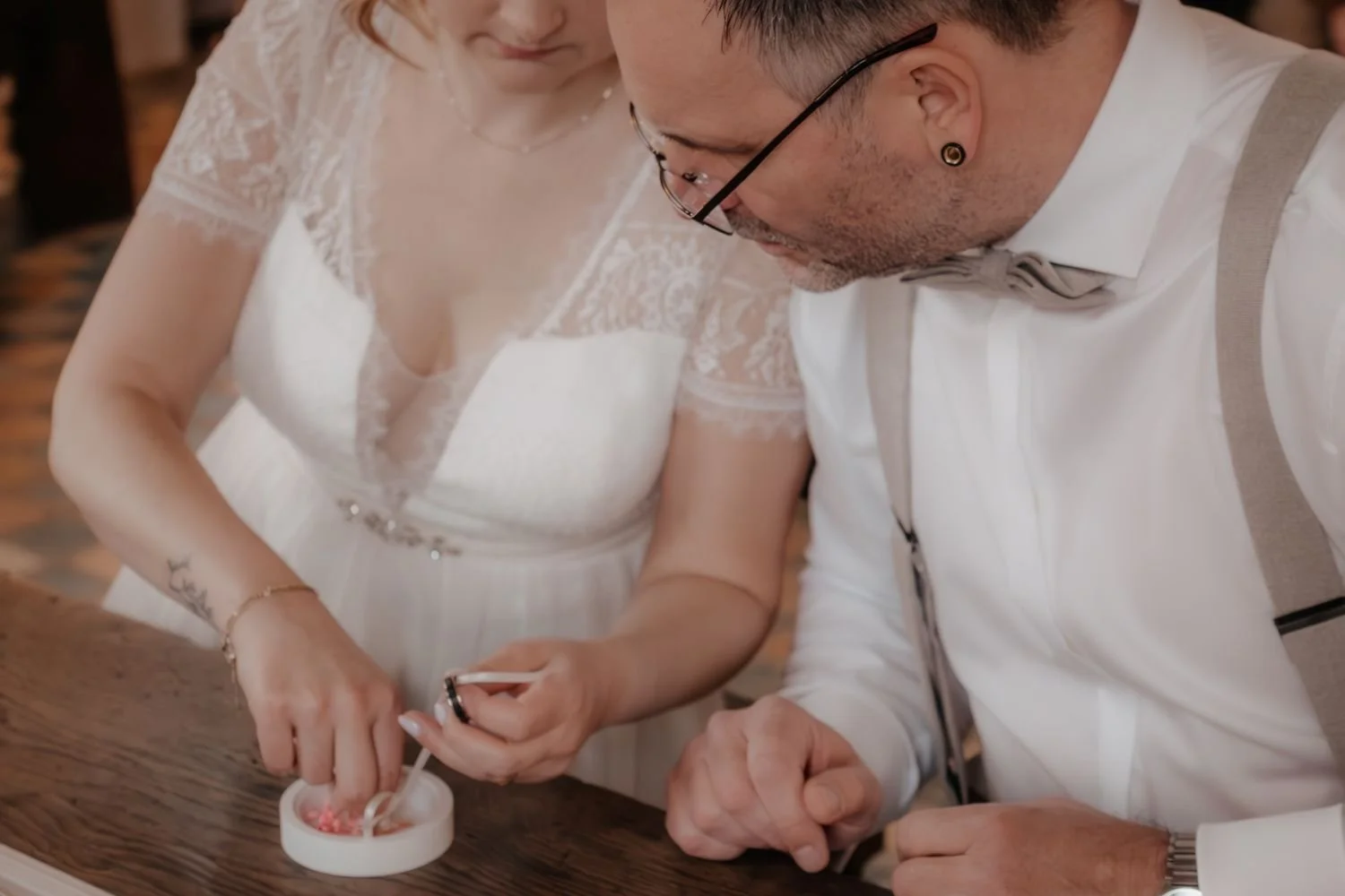 Ein Paar in Hochzeitskleidung bei einer Zeremonie, sie schauen auf einen Ring, der über ein kleines Batteriefässchen mit einem Zettel gehalten wird.