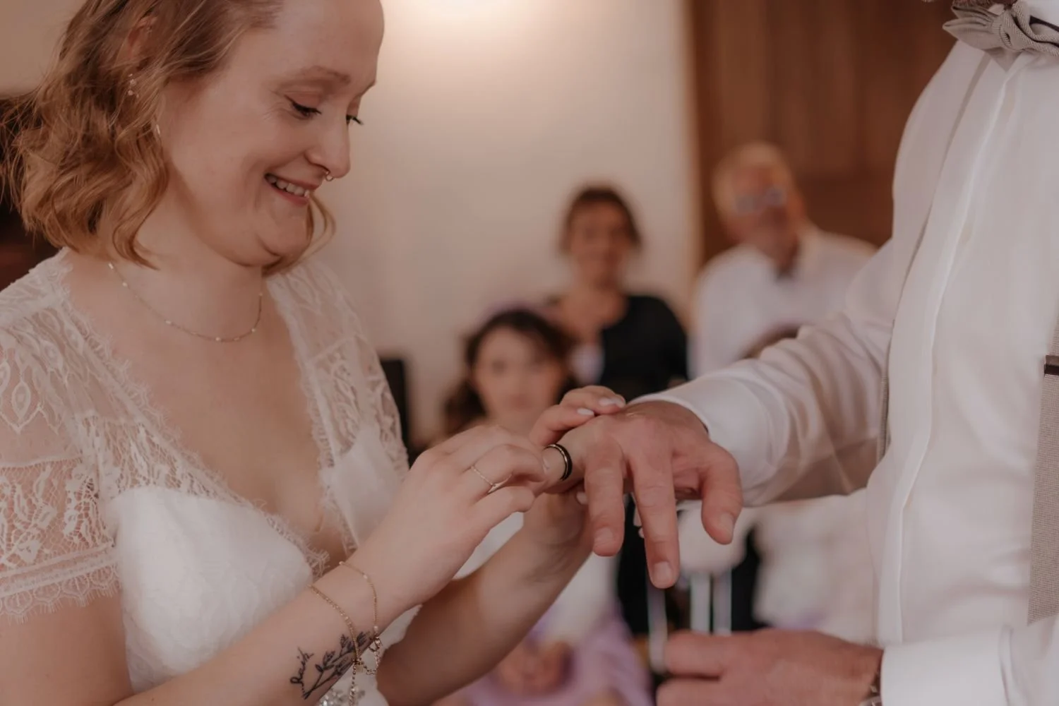 Ein Brautpaar tauscht Eheringe bei einer Hochzeit, die Braut lächelt, im Hintergrund sind Gäste zu sehen.