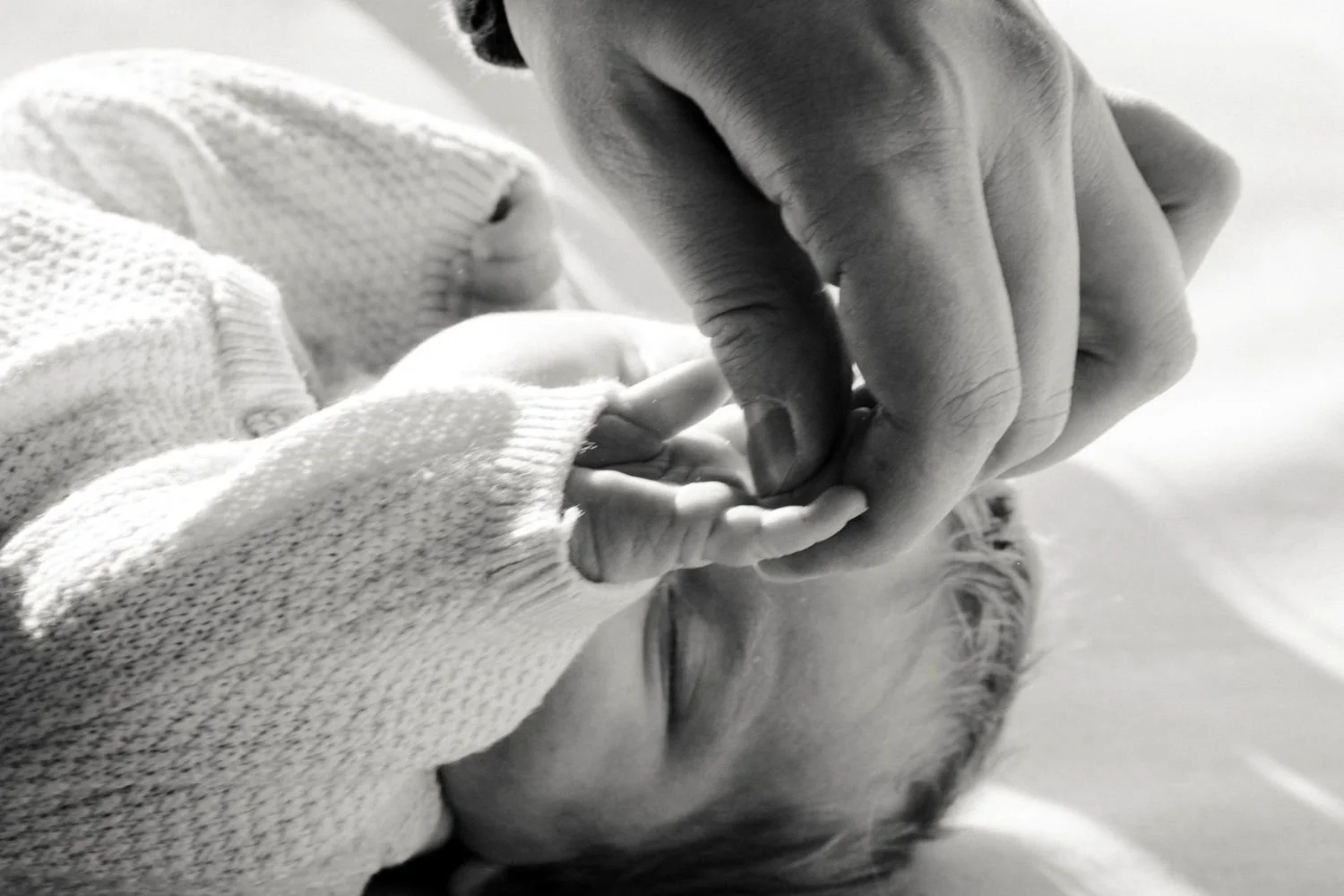 Ein Baby liegt auf dem Rücken und hält die Hand des Papas, während der Papa die Hand des Kindes berührt. Das Foto ist in Schwarz-Weiß.