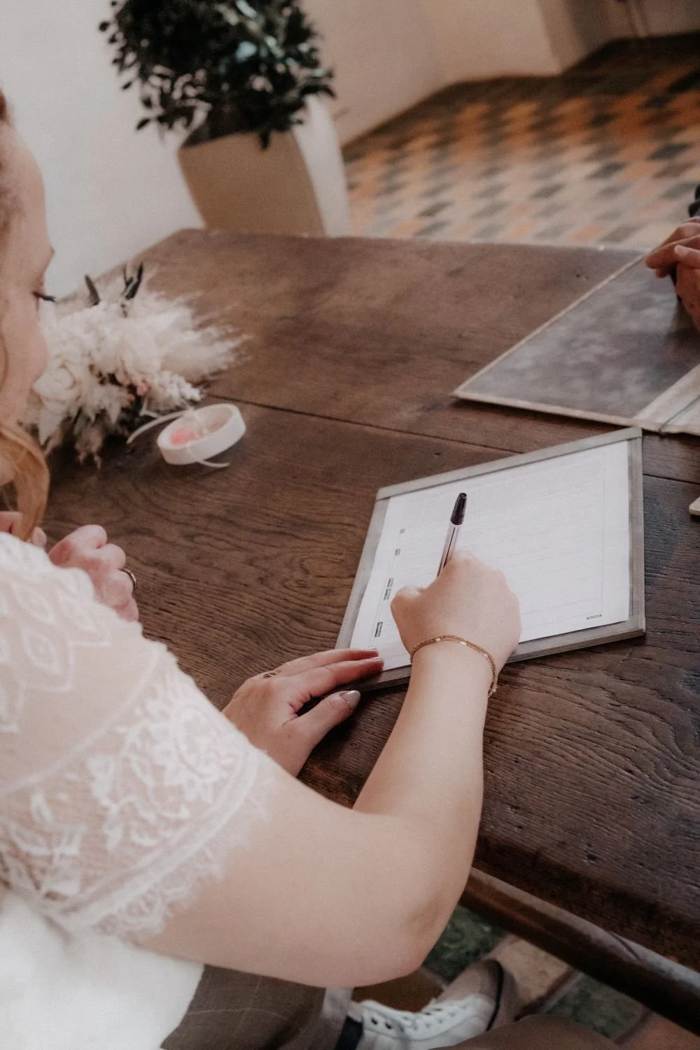 Eine Frau schreibt auf einem Notizblock, während sie an einem Holztisch sitzt. Im Hintergrund ist ein Büro oder Café mit Fliesenboden und einer Pflanze zu sehen.