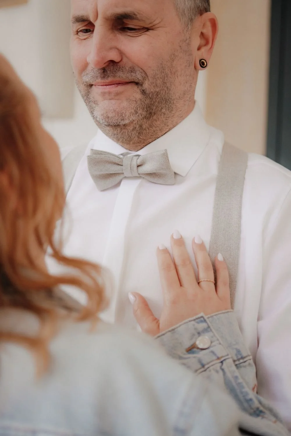 Ein Mann trägt einen weißen Hemd mit einer grauen Fliege und Hosenträger, eine Frau berührt seine Brust. Es ist eine emotionale Szene, vermutlich bei einer Hochzeit.