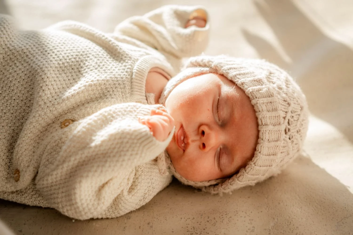Ein schlafendes Baby in einem weißen gestrickten Outfit mit Mütze, liegend auf einer weichen Oberfläche.