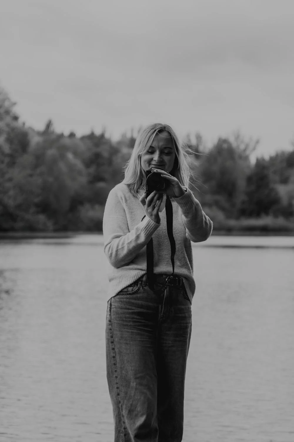 Junge Frau in Freizeitkleidung steht an einem Fluss, fotografiert sich selbst mit einer Kamera, im Hintergrund Bäume, in Schwarz-Weiß.