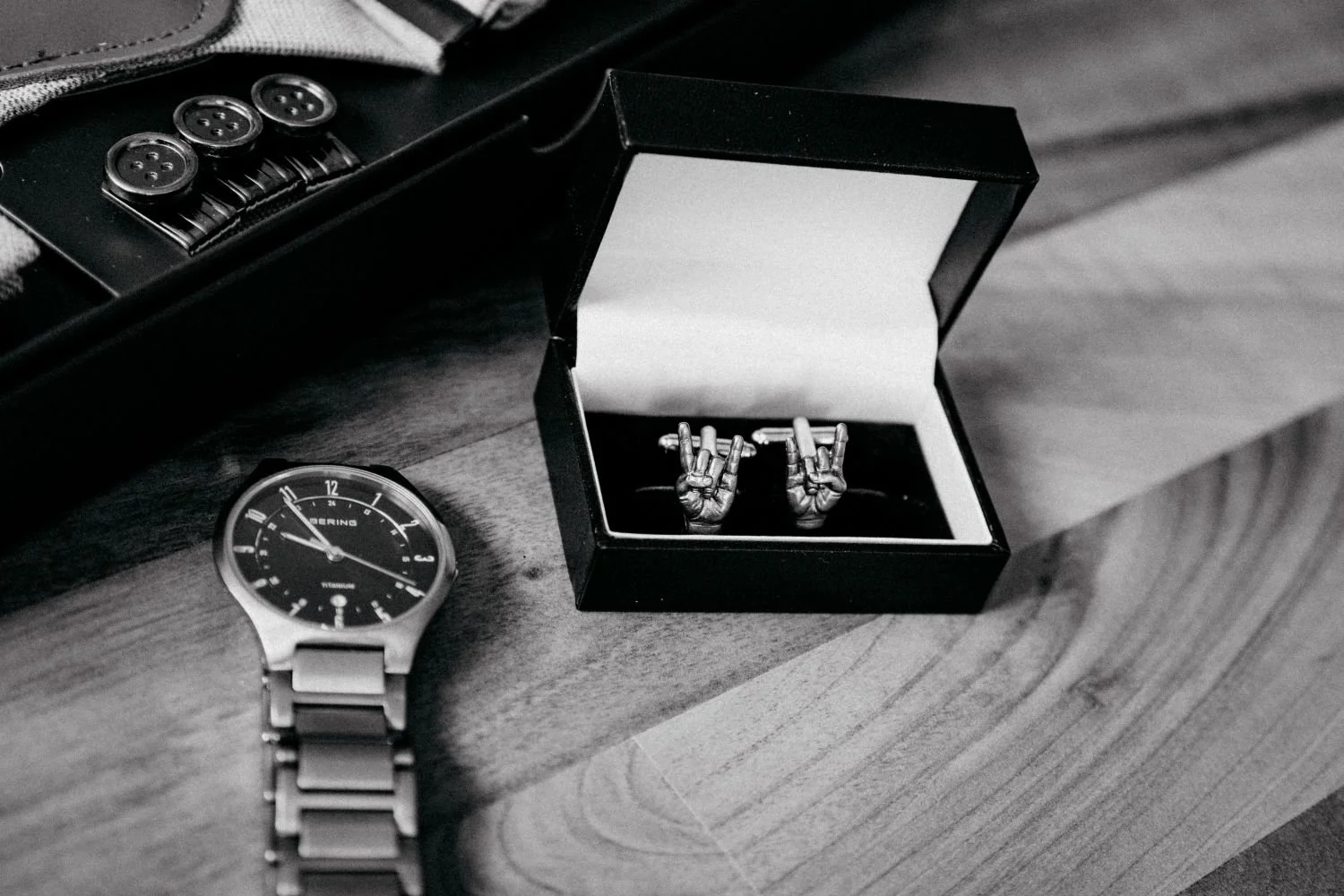 Eine Armbanduhr, ein offenes Geschenkbox mit einer Manschettenknopf-Weiterentwicklung in Form von Handschuhen, und ein Taschenmesser auf einem Holztisch.