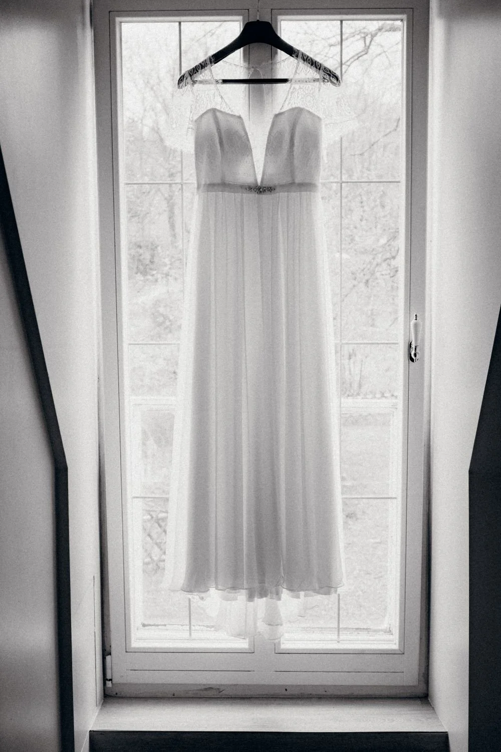 Ein weißes Brautkleid hängt an einem Kleiderbügel vor einem Fenster. Das Kleid ist bodenlang mit Spaghettiträgern und floralen Details an den Schultern.