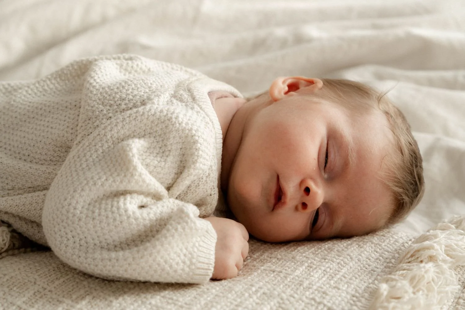 Kleiner Junge schläft auf einem weißen Bett, eingekuschelt in eine cremefarbene Strickjacke.