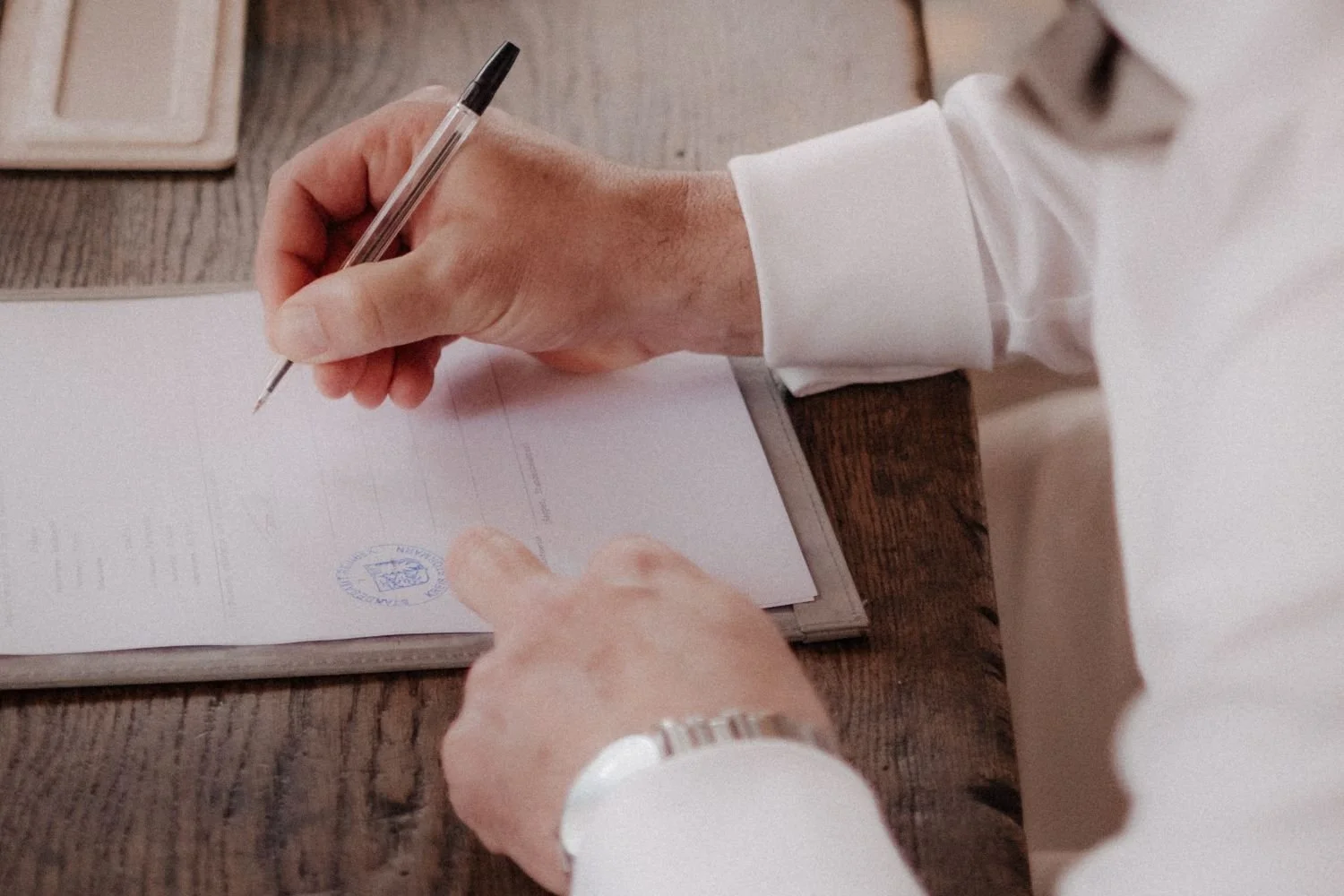 Hand eines Mannes in Businesskleidung unterschreibt ein Dokument mit einem Kugelschreiber auf einem Holztisch.