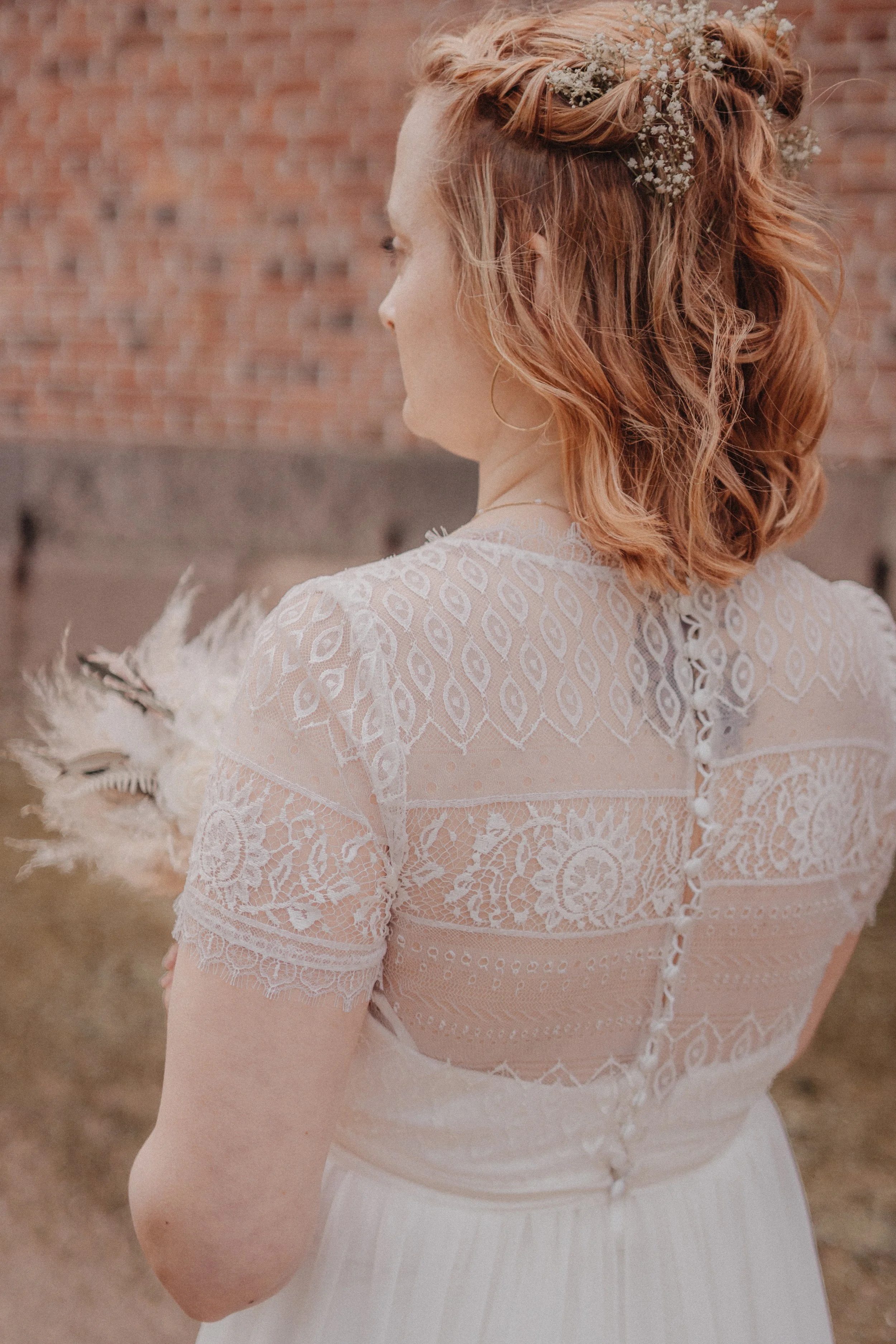 Eine Frau in einem weißen Brautkleid mit Spitze, mit roten, lockigen Haaren und Blumen im Haar, steht vor einer Backsteinmauer.