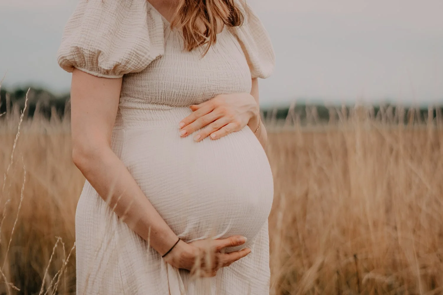 Eine schwangere Frau, die ihren Bauch in einem Weizenfeld hält, trägt ein helles Kleid mit puffigen Ärmeln.