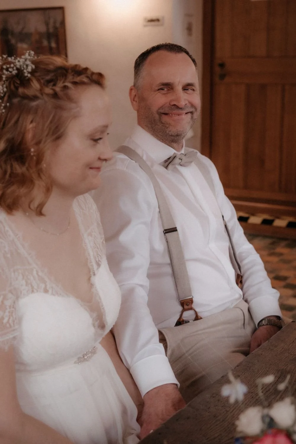 Ein Mann und eine Frau sitzen an einem Tisch bei einer Hochzeit, der Mann lächelt und trägt ein weißes Hemd mit Hosenträgern und Schleife, die Frau trägt ein weißes Hochzeitskleid mit Spitze und einen Haarreif mit Blumen.