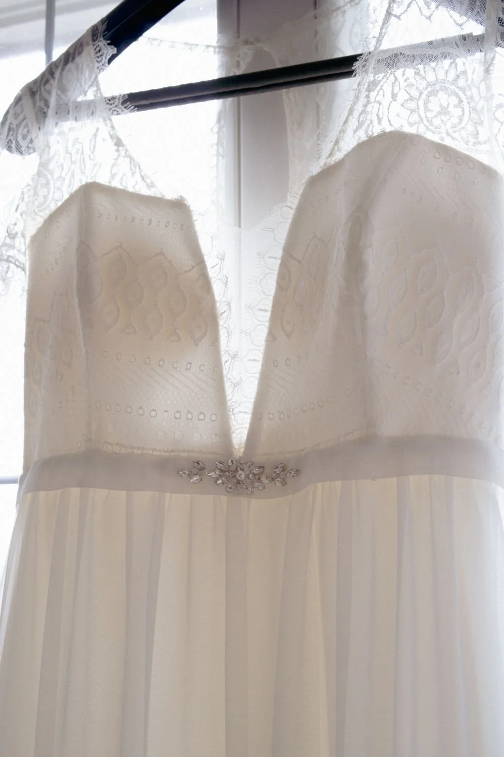 Weiße Hochzeitsbrautkleid mit Spitzenärmeln, Perlen und Schmuckbesatz, auf einem Bügel vor einem Fenster aufgehängt.