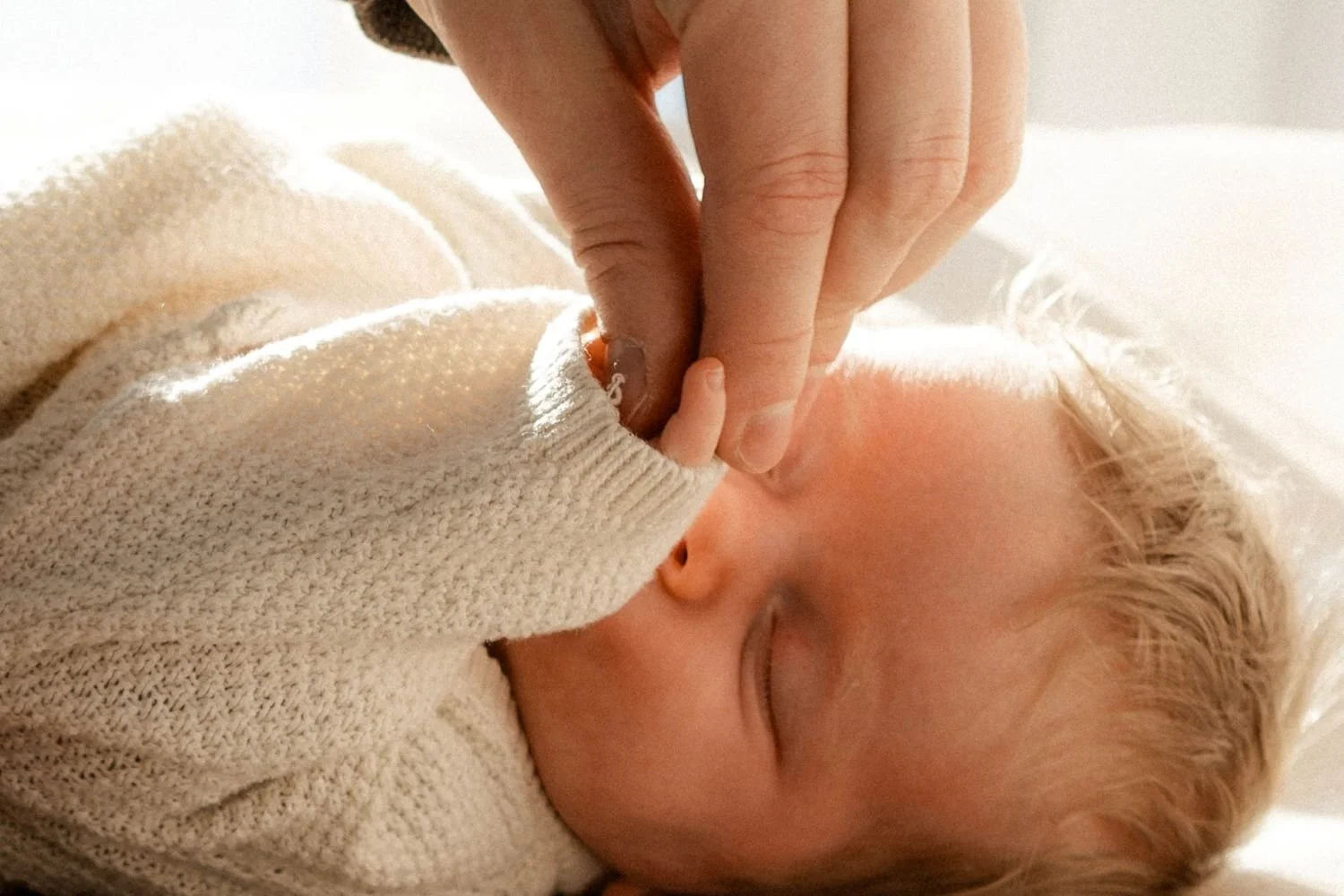 Ein neugeborenes Baby schläft friedlich und jemand berührt liebevoll seine Hand. Das Baby trägt einen hellen, gestrickten Pullover und liegt auf weißen Bettlaken.