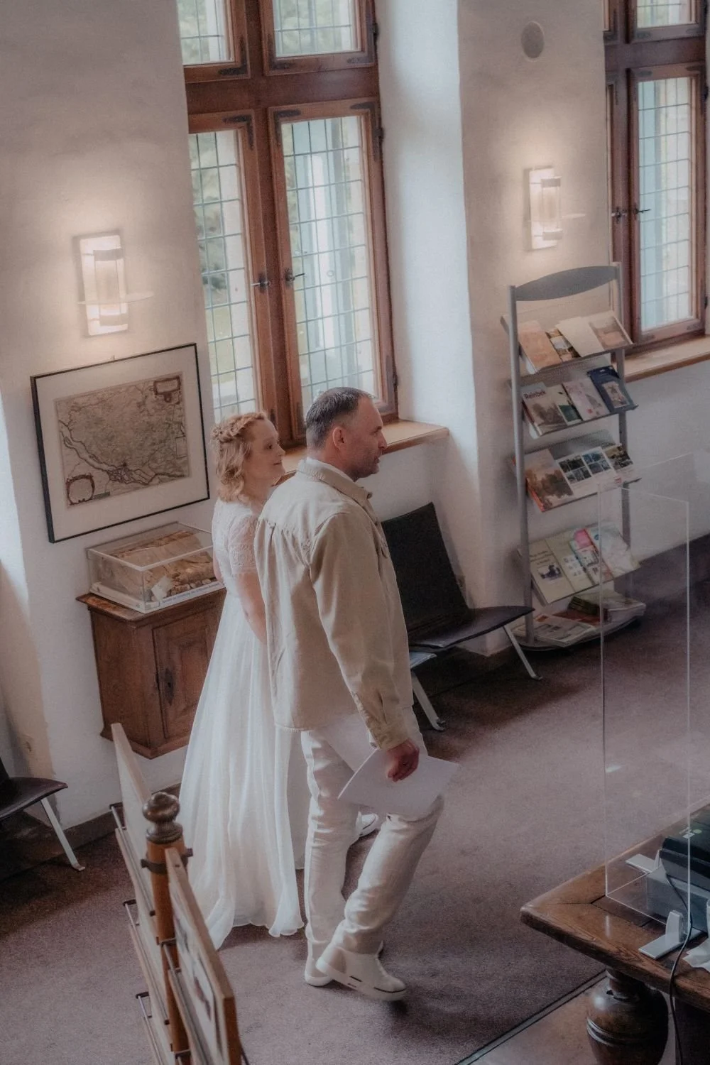 Ein Mann und eine Frau stehen in einem Raum mit alten, großen Fenstern. Die Frau trägt ein weißes Kleid, der Mann ein helles Jackett. Der Raum ist mit Büchern und Bildern dekoriert.