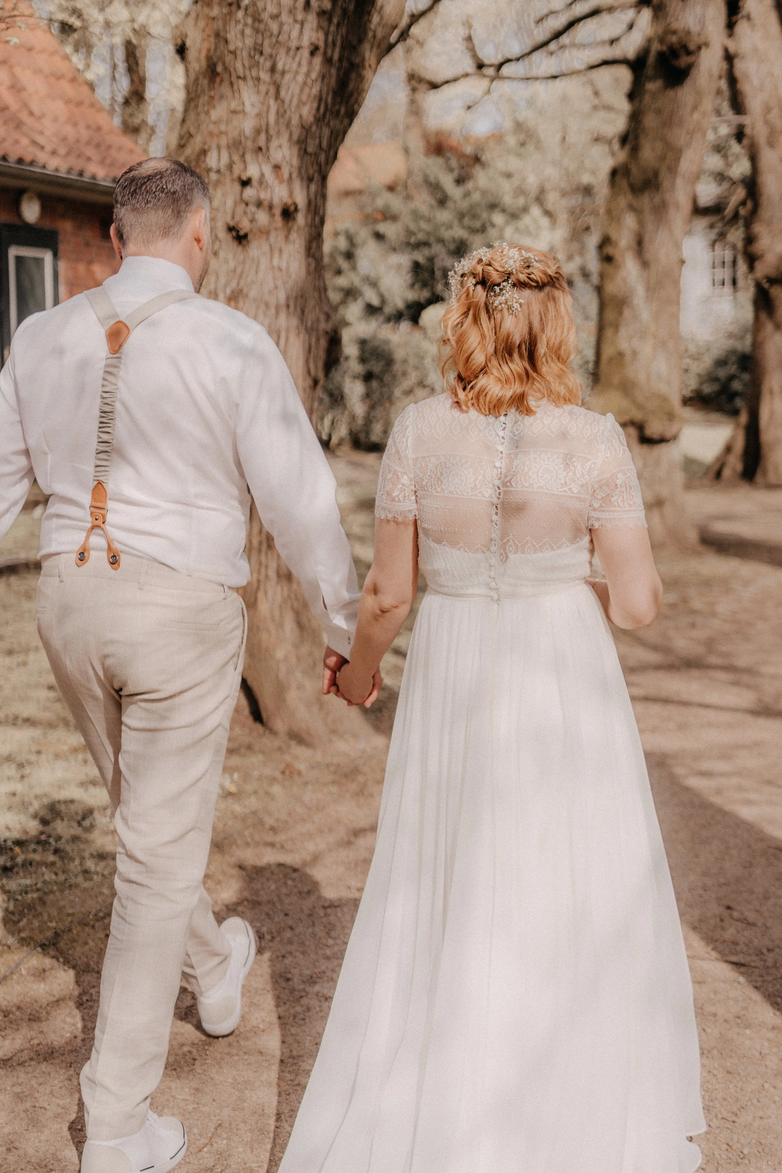 Ein Brautpaar hält Händchen, während es durch einen parkähnlichen Bereich mit großen Bäumen geht. Das Mädchen trägt ein langes weißes Hochzeitskleid mit Spitze, der Junge trägt beige Hosen und ein weißes Hemd mit Hosenträgern.