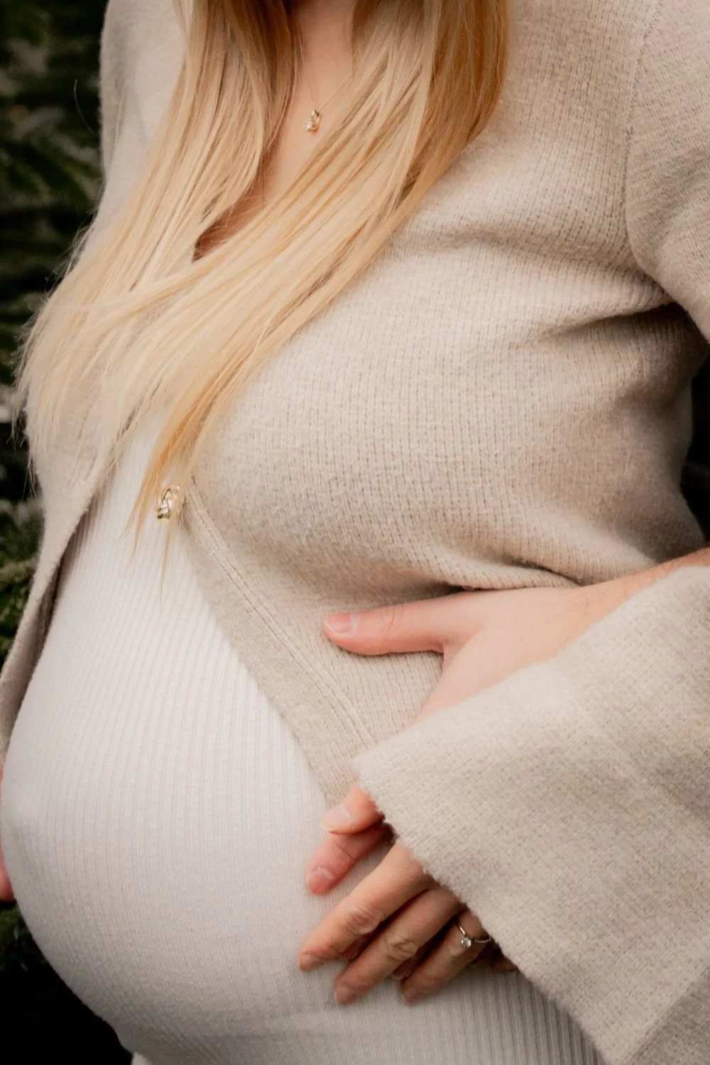 Nahaufnahme eines schwangeren Bauches, gehalten von einer Hand, mit Frauenarm in einem hellen Pullover im Hintergrund, im Freien.