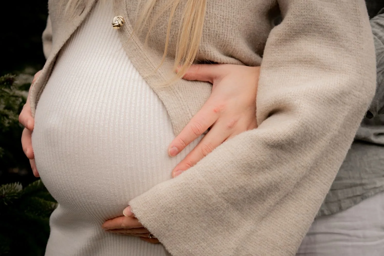Eine schwangere Frau hält ihre Bauch mit beiden Händen, trägt einen beigen Mantel und eine weiße Hose.