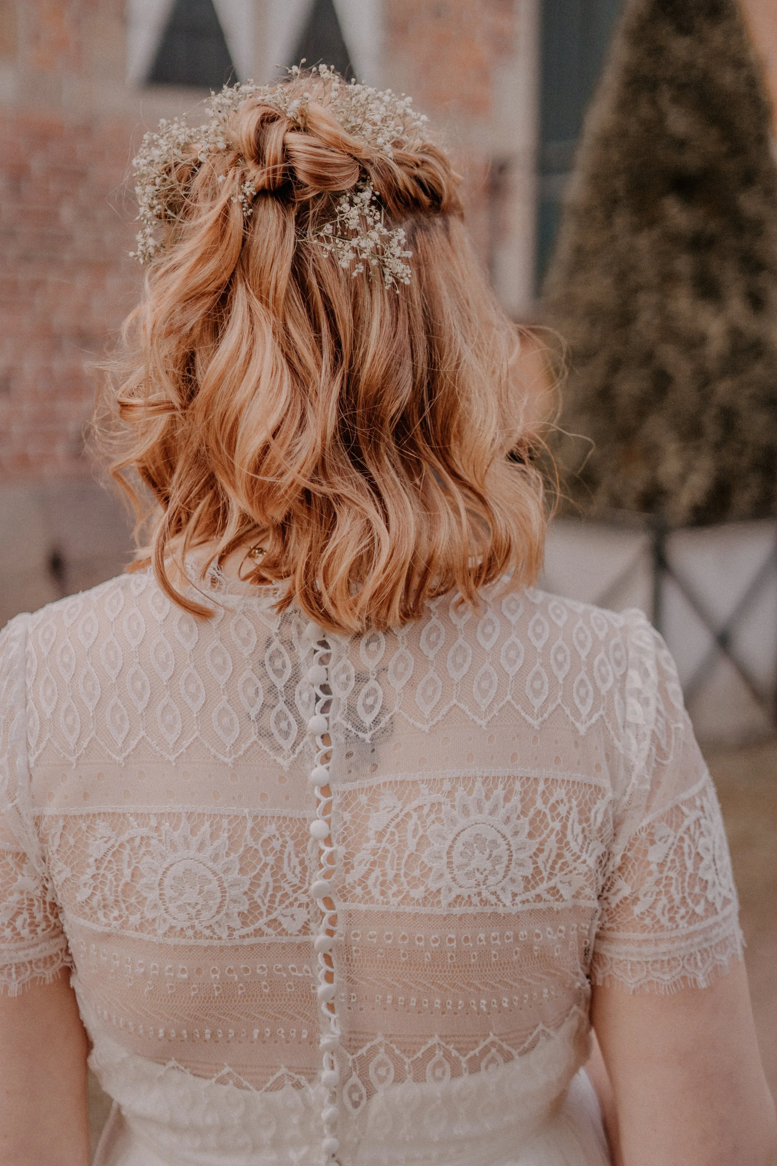 Eine Frau mit lockigem, rotem Haar, die eine weiße Spitzenkleidung trägt und eine kleine Blumenkrone auf dem Kopf hat, steht im Freien vor einem Backsteinmauer und Bäumen.