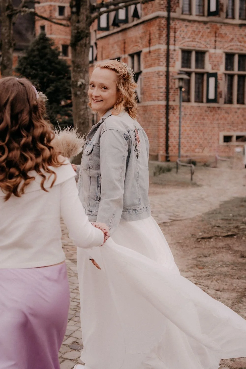 Zwei Mädchen, eines in einem weißen Kleid und das andere in einem grauen Jacke, halten die Hände und lachen vor einem Backsteingebäude im Freien.