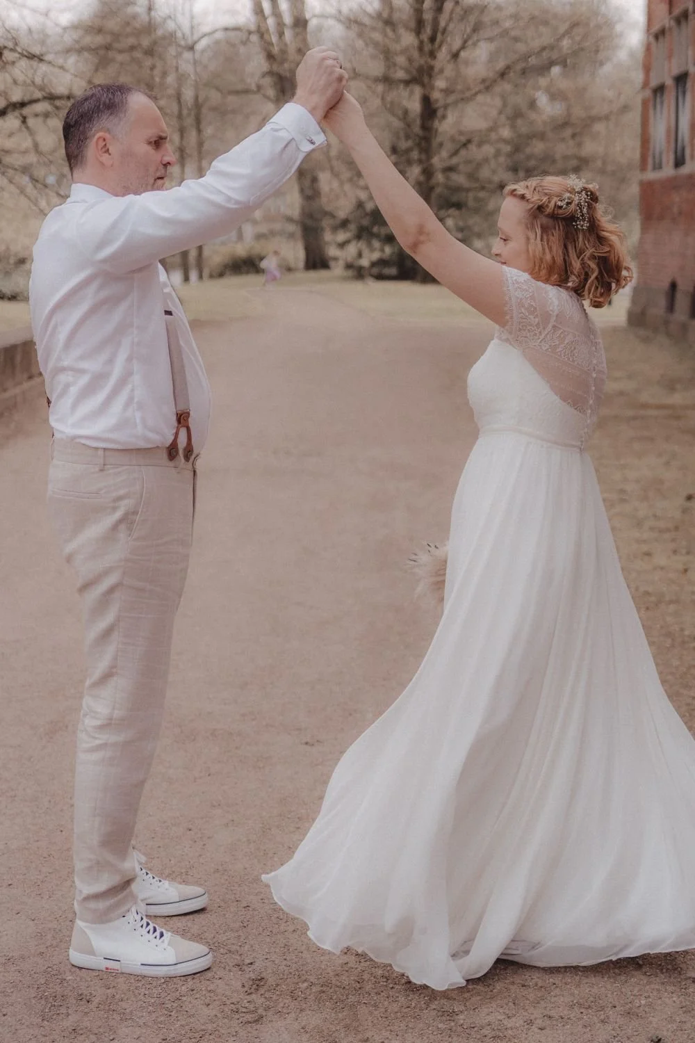 Ein Mann und eine Frau tanzen im Freien, die Frau trägt ein weißes Brautkleid, der Mann eine weiße Bluse und helle Hosen, im Hintergrund sind Bäume und eine rote Backsteinbaut. Es sieht nach einer Hochzeit aus.