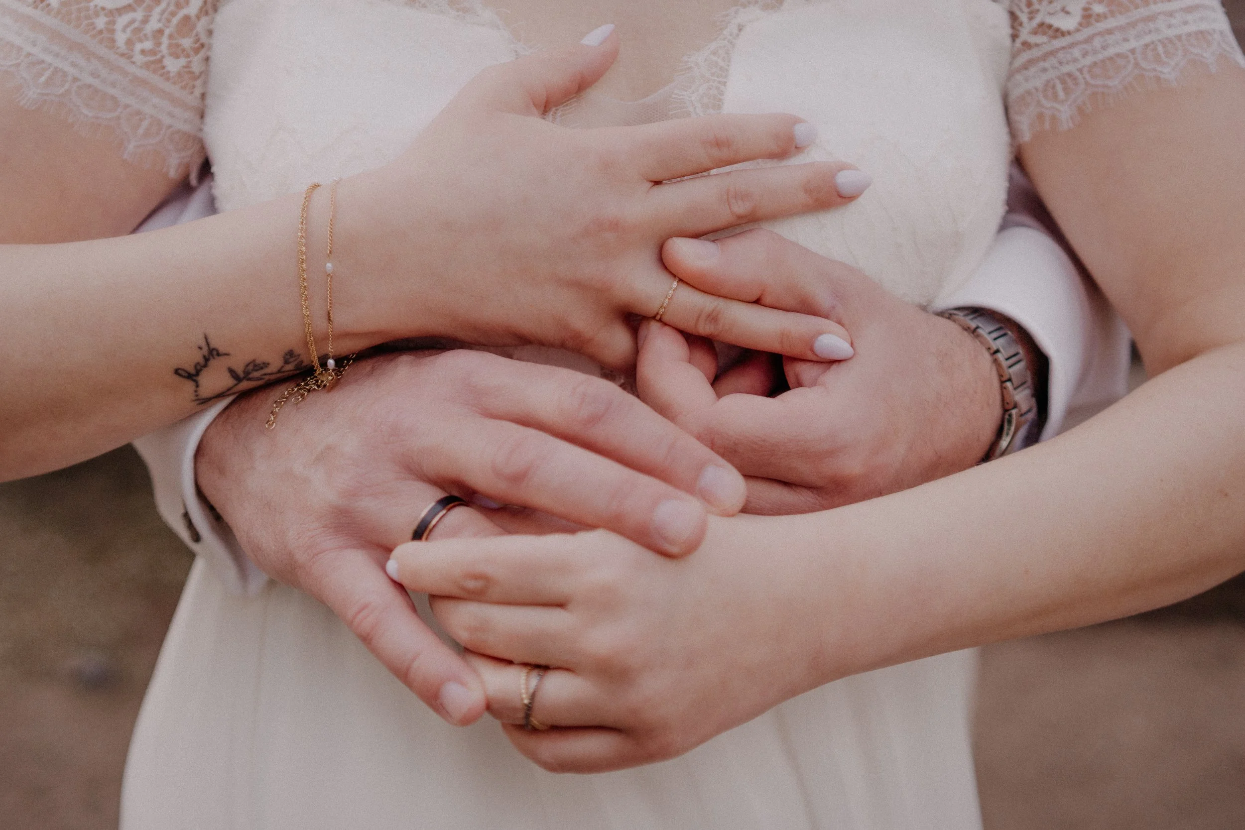 Nahaufnahme von vier Händen, die sich auf einem weißen Kleid berühren. Zwei Hände gehören einer Frau mit weißen Nagellack, zartem Schmuck und Tattoos, die anderen beiden einer Person in einem weißen Hemd mit Uhr und Ring. Es handelt sich wahrscheinli