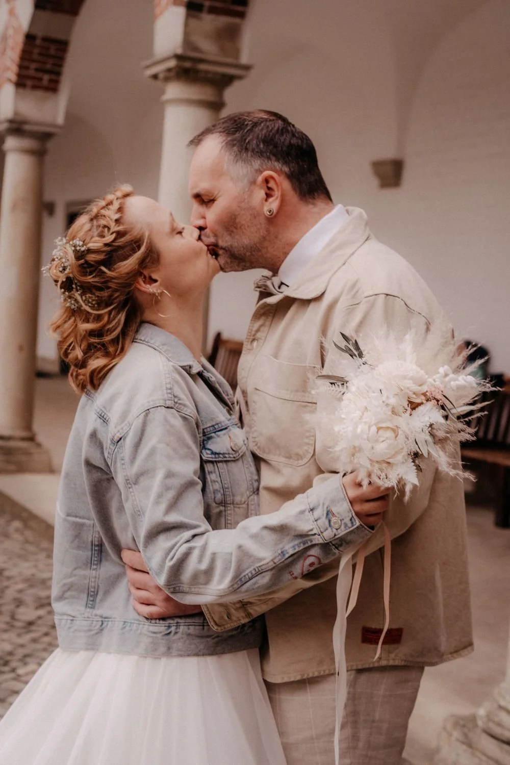 Ein Paar küsst sich bei einer Hochzeit, die Frau trägt ein Brautkleid und eine Jacquardjacke, der Mann trägt einen beigen Sakko und hält einen Brautstrauß aus weißen Blumen. Sie stehen in einer alten Kirche mit Säulen im Hintergrund.